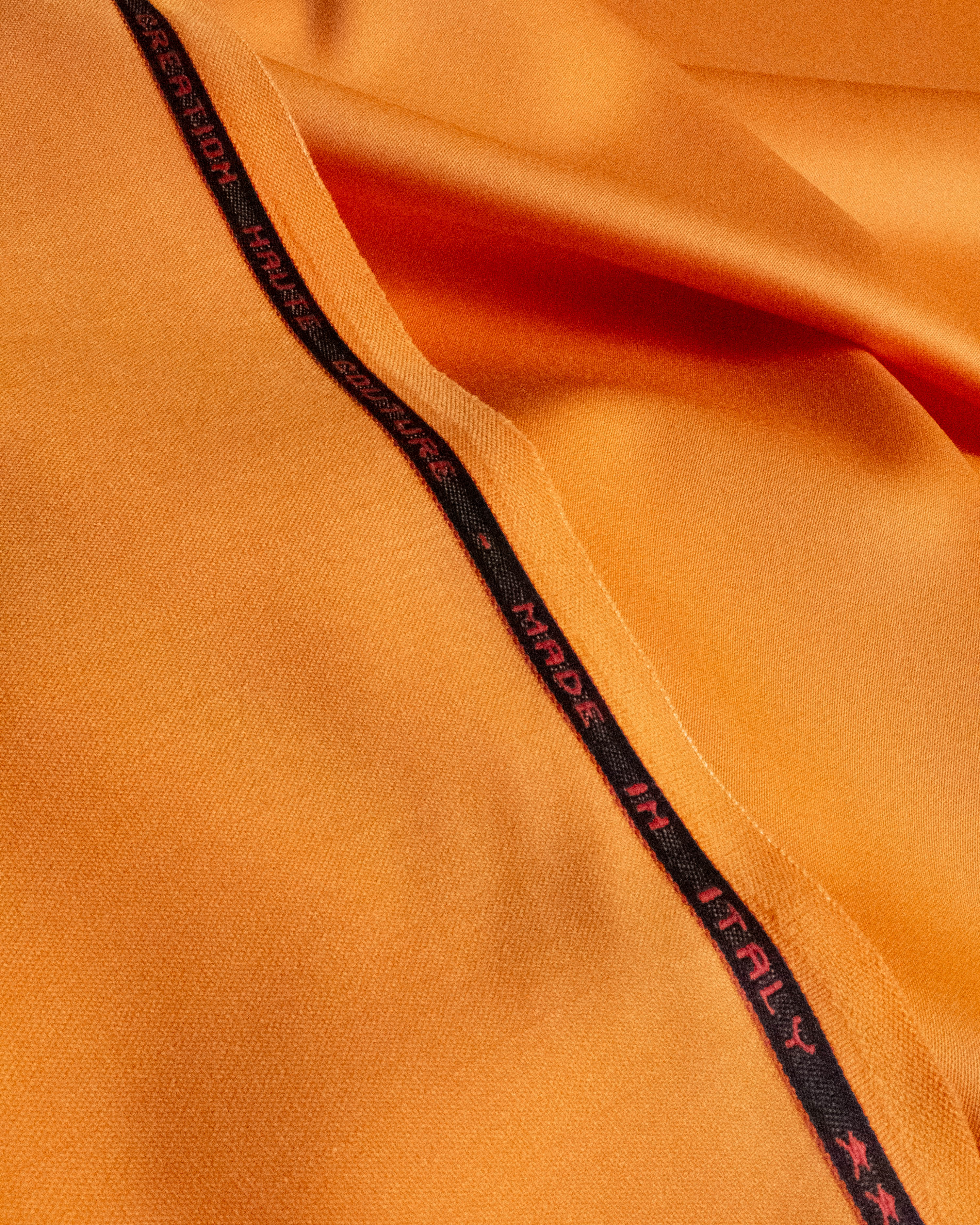 Close-up of a bright orange fabric with a black branded trim running diagonally across the frame. The trim features repeated red text reading 'MADE IN ITALY' alongside the Puma cat logo.
