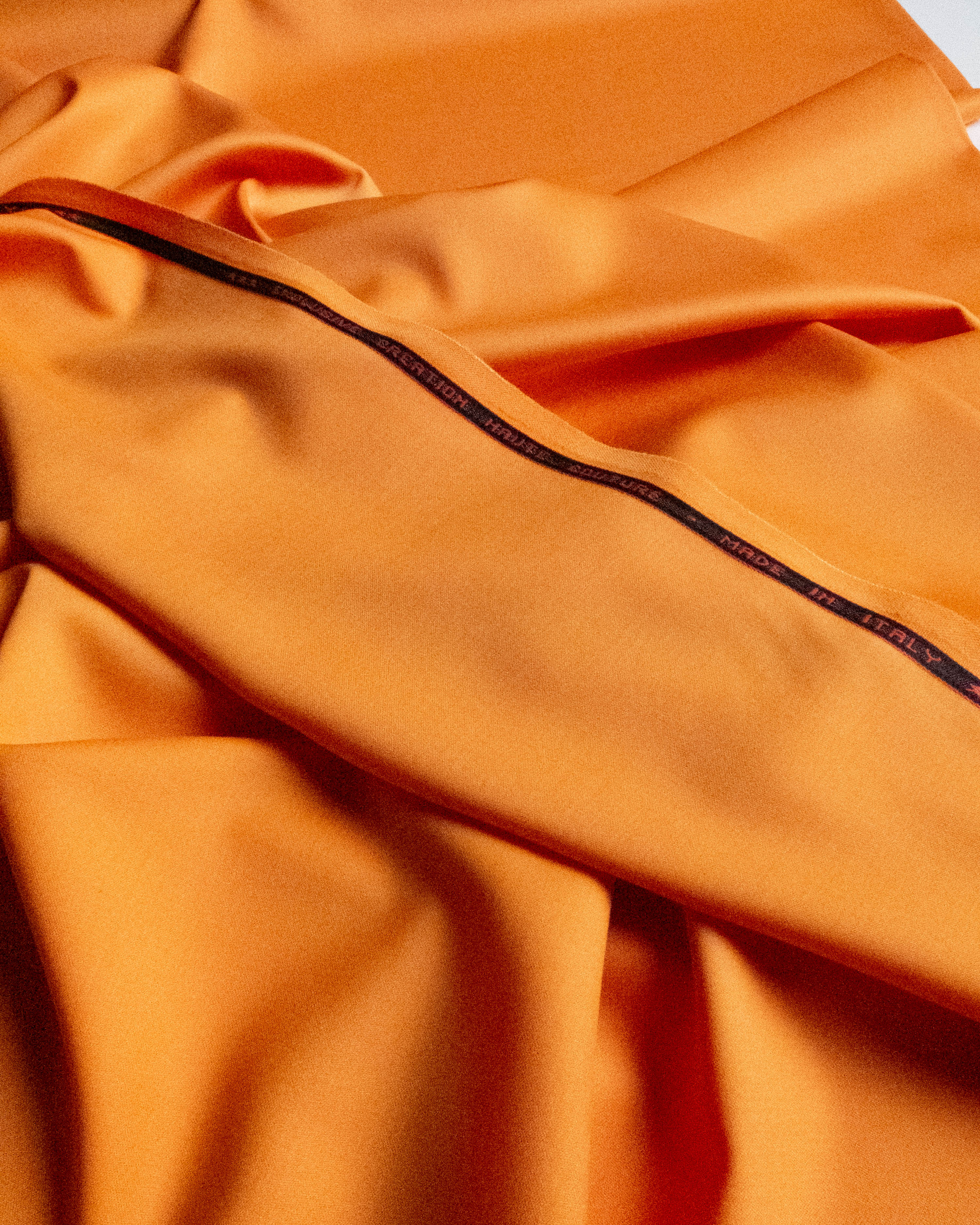 A close-up of draped orange satin fabric with a thin black stitched trim running diagonally across the surface.