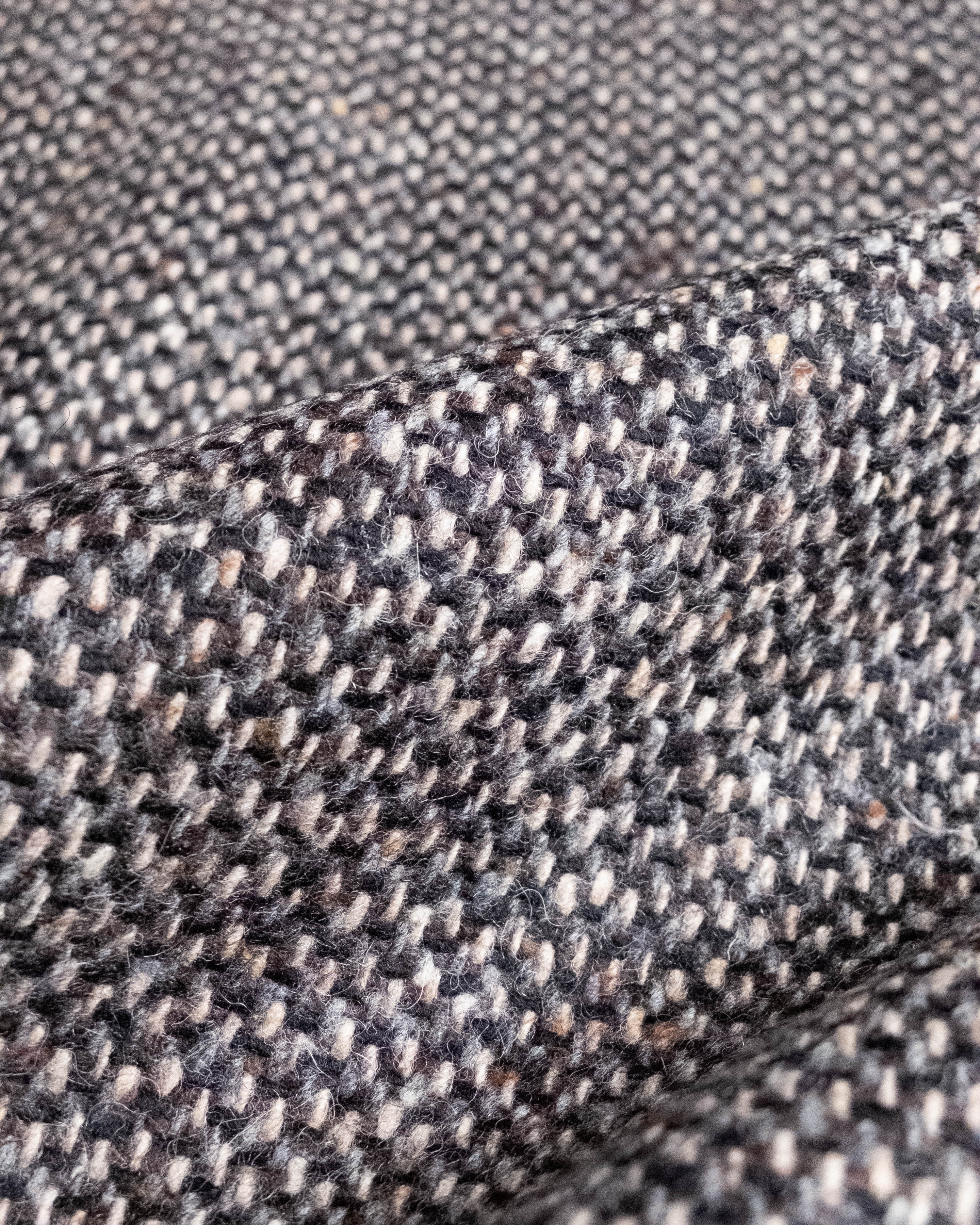 Close-up of a textured woven fabric with a herringbone pattern in dark gray, black, and beige threads, showing the detailed weave and soft fold.