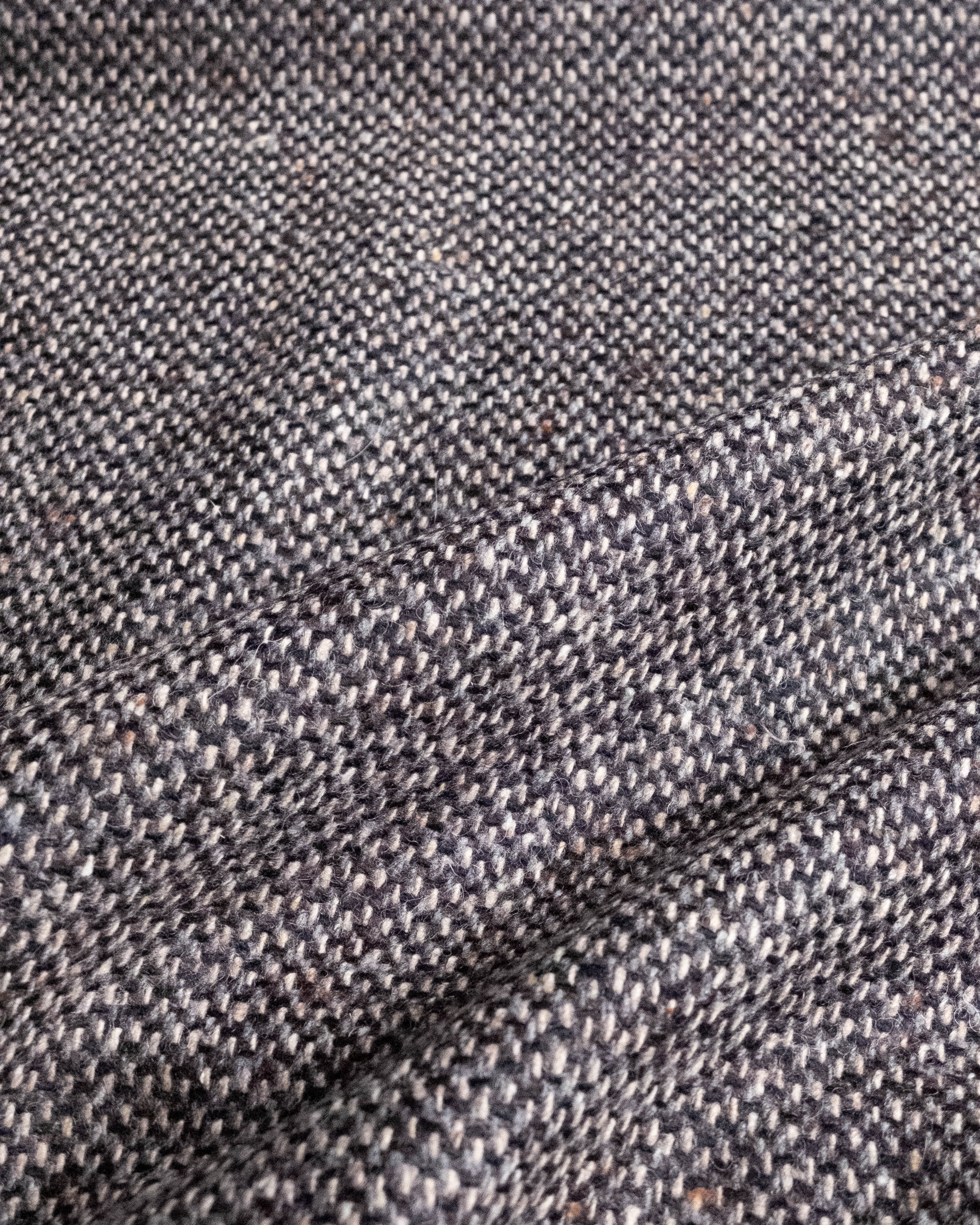 A close-up of a textured woven fabric with a heathered pattern of gray, black, and white threads, showing a fine tweed or herringbone weave.