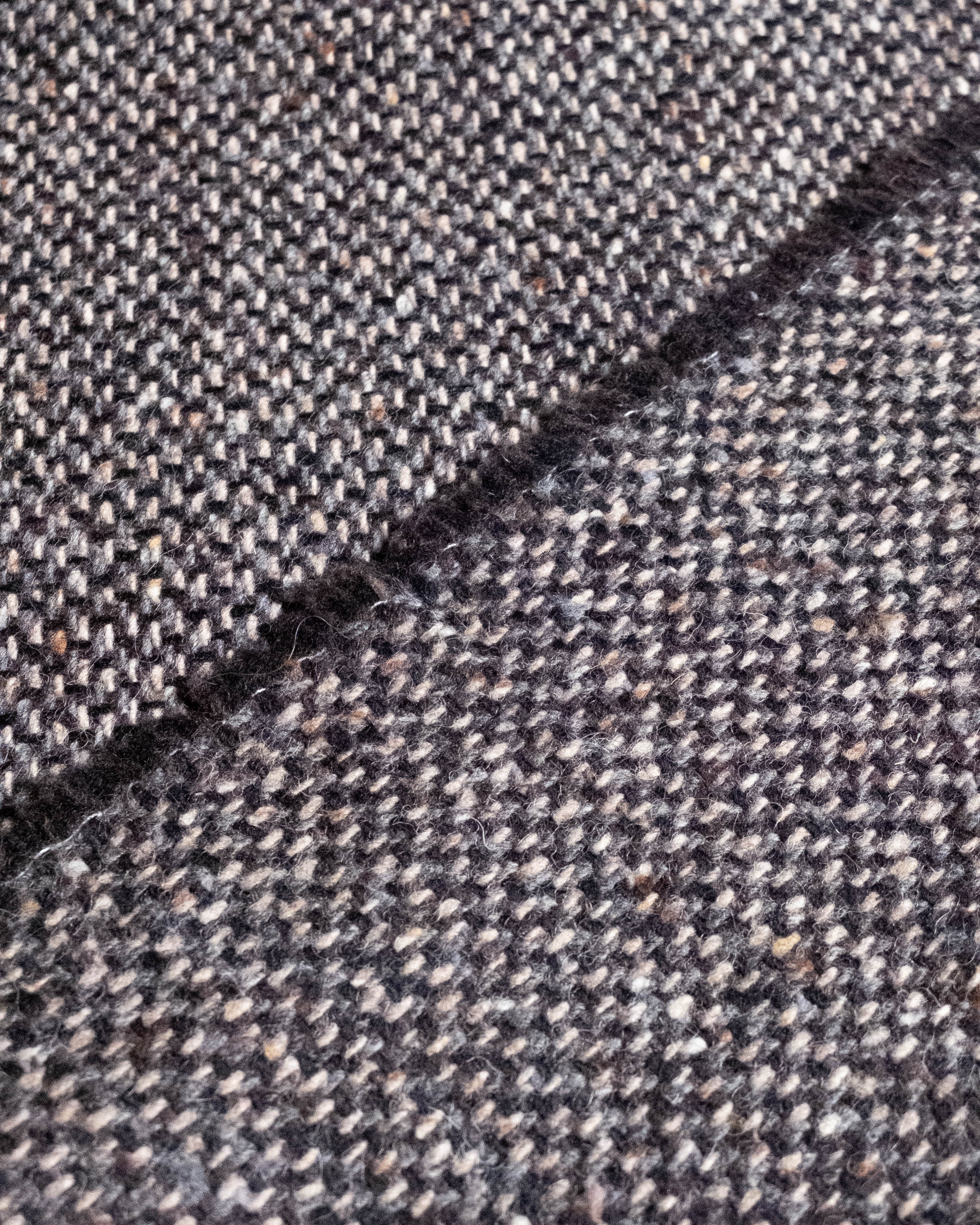 Close-up of a textured tweed fabric with a diagonal fold, showing a mottled pattern of dark gray, brown, and off-white threads.